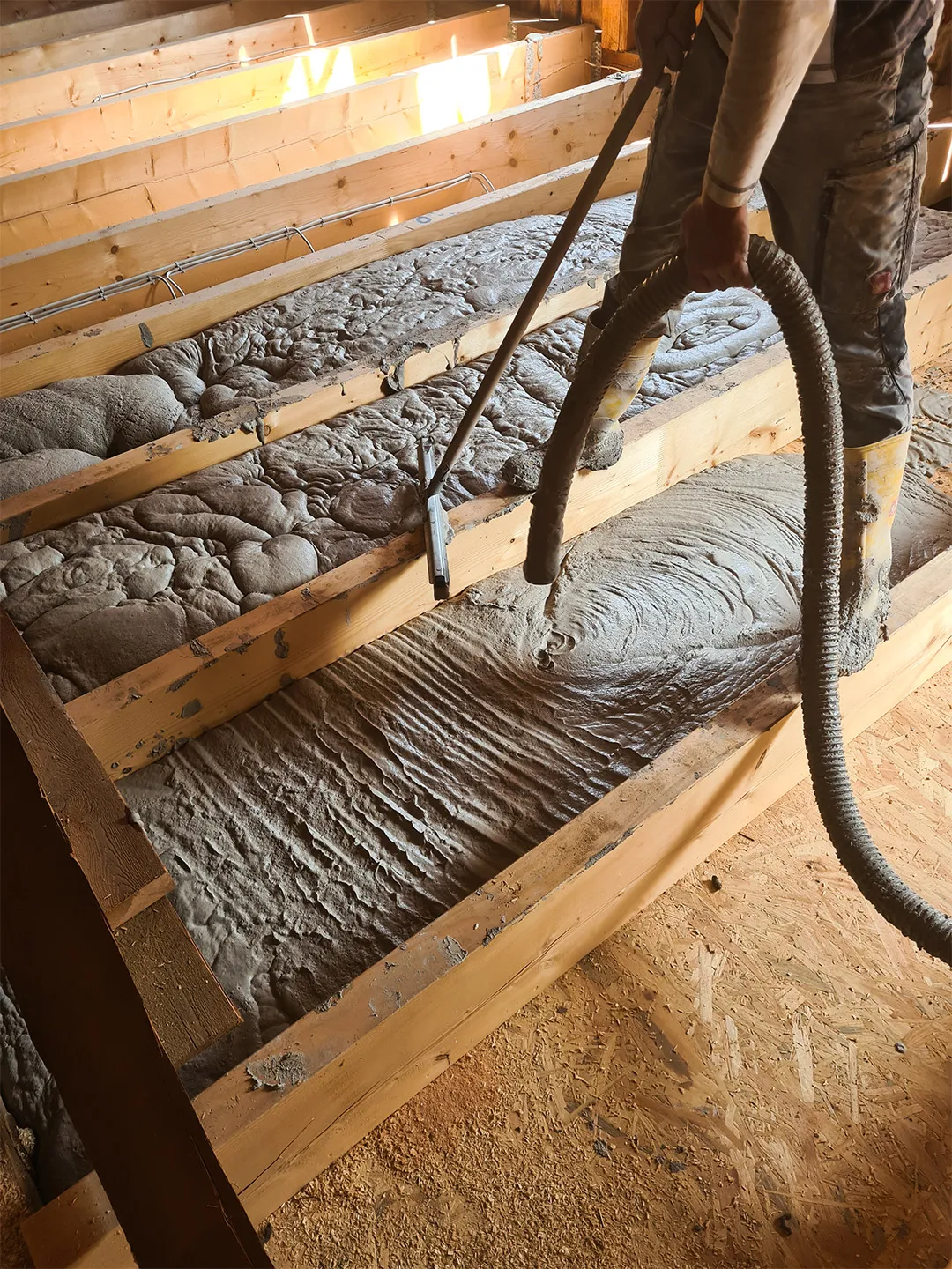 Ein Bodenaufbau mit sichtbaren Holzbalken, die mit frischer Schüttdämmung befüllt werden, ein Arbeiter mit einem Schlauchwerkzeug im Einsatz