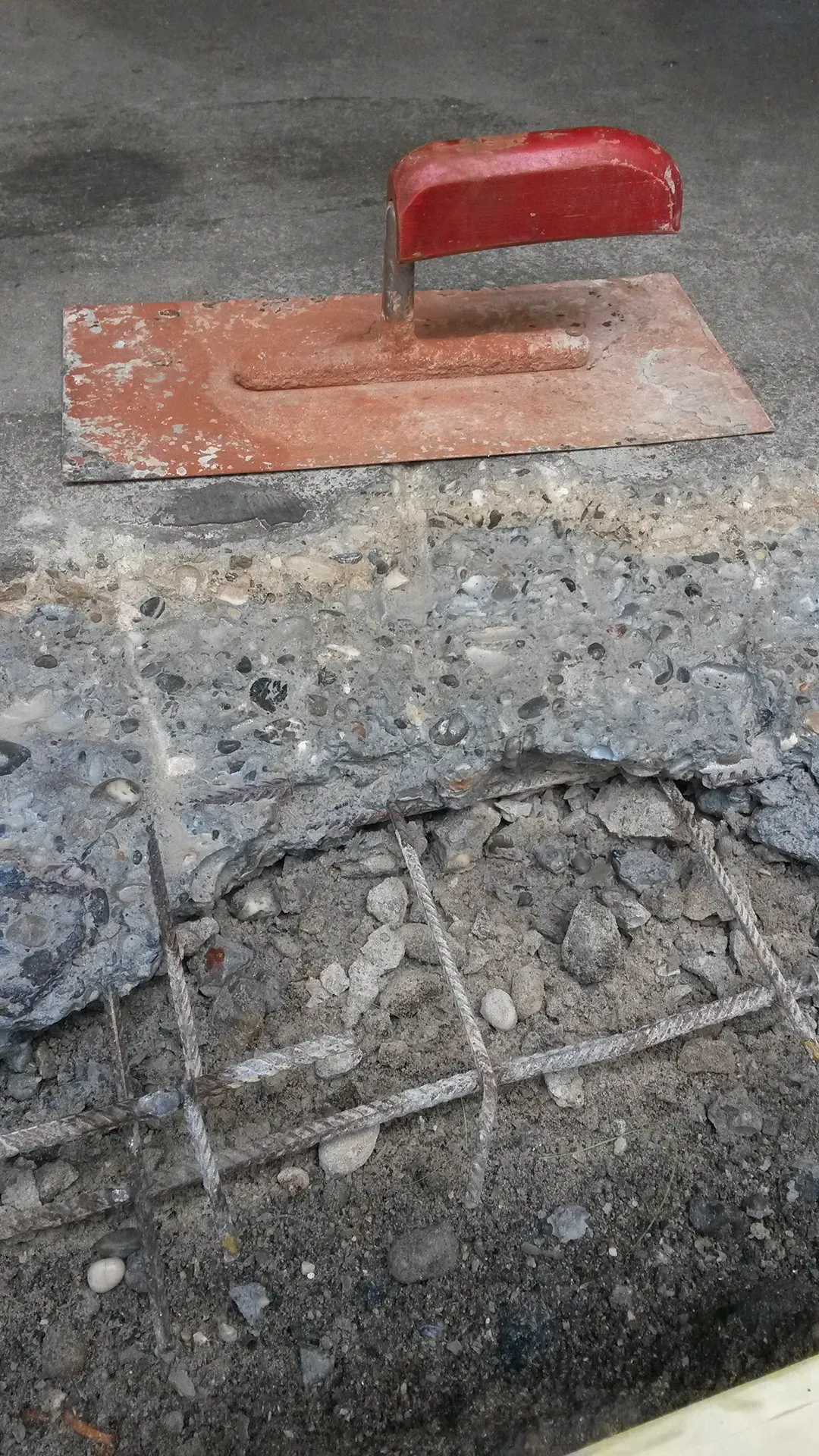Nahaufnahme eines roten Handglätters auf herausgebrochenem Beton mit freiliegender Bewehrung