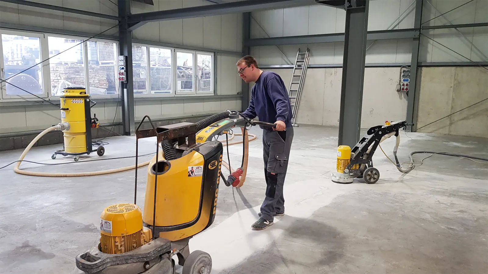 Werkshalle mit Schleifmaschinen für Betonböden, ein Mitarbeiter führt Schleifarbeiten durch
