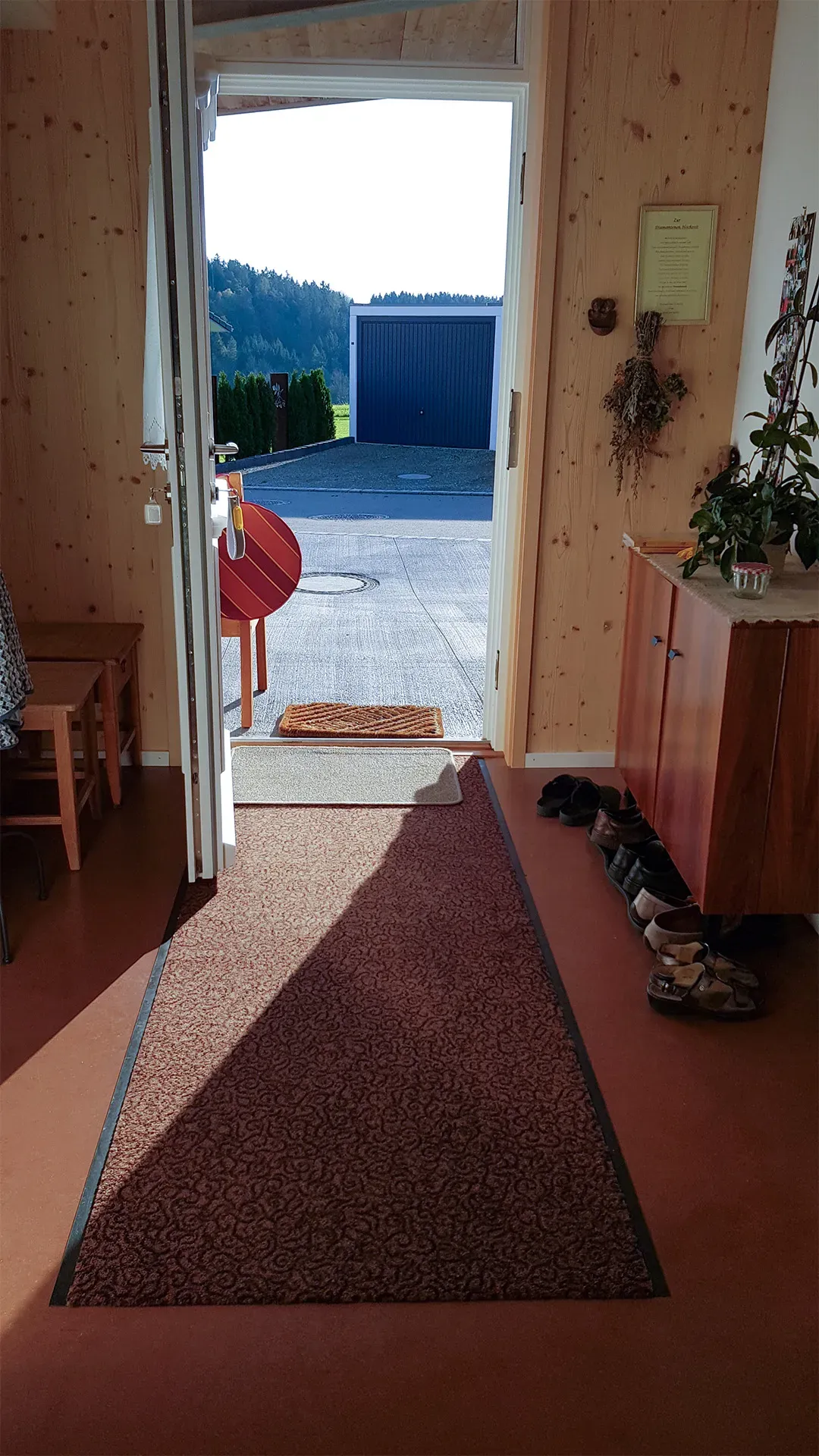 Offene Haustür eines Hauses mit Blick ins Freie, ein roter Teppich im Eingangsbereich und Schuhe am Rand